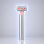 A sleek, modern LED light therapy wand with a brushed silver metal body, a soft pink accent collar, and a silver T-shaped head featuring a glowing red wavy LED light. It stands vertically on a reflective surface against a clean, light gray gradient background.